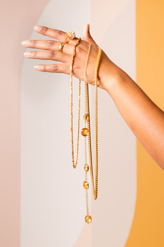 A hand draped with layered gold chain necklaces against a warm orange and cream backdrop