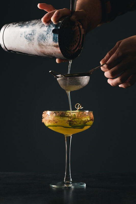 A bartender straining a yellow cocktail into a coupe glass against dark slate