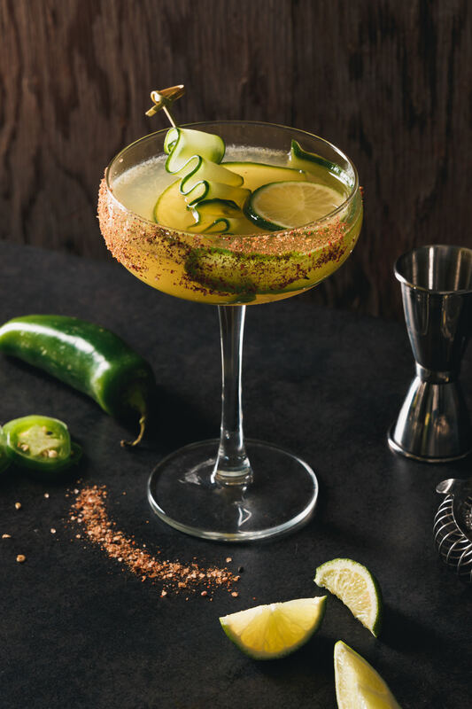 A cucumber and lime cocktail in a coupe glass with chili and spices on a dark table