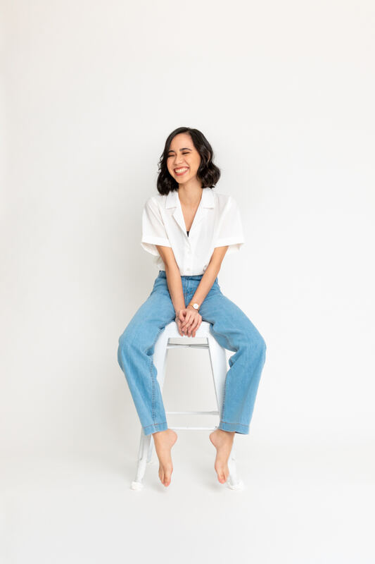A woman in a white shirt and jeans seated on a stool against a white backdrop