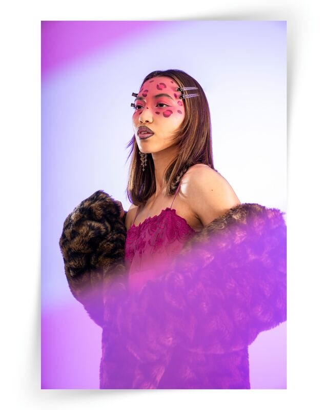 A model in a magenta feathered outfit and pink sunglasses under violet light