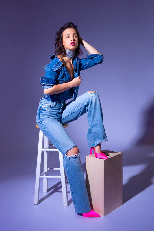 A model in denim and pink heels perched on a stool under violet light