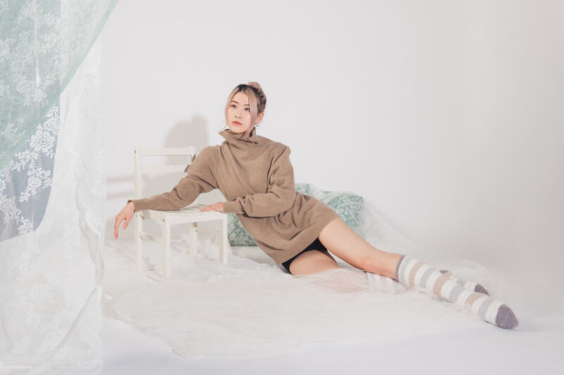 A model in a camel coat lounging on a white bed by sheer curtains